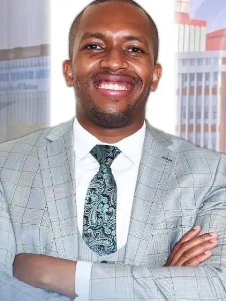 Rev. Elvis Mathenge smiling, in a grey checked suit, arms folded
